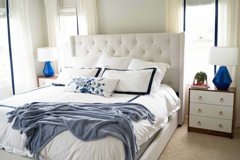 upholstered king size bed with white sheets in front of windows master bedroom