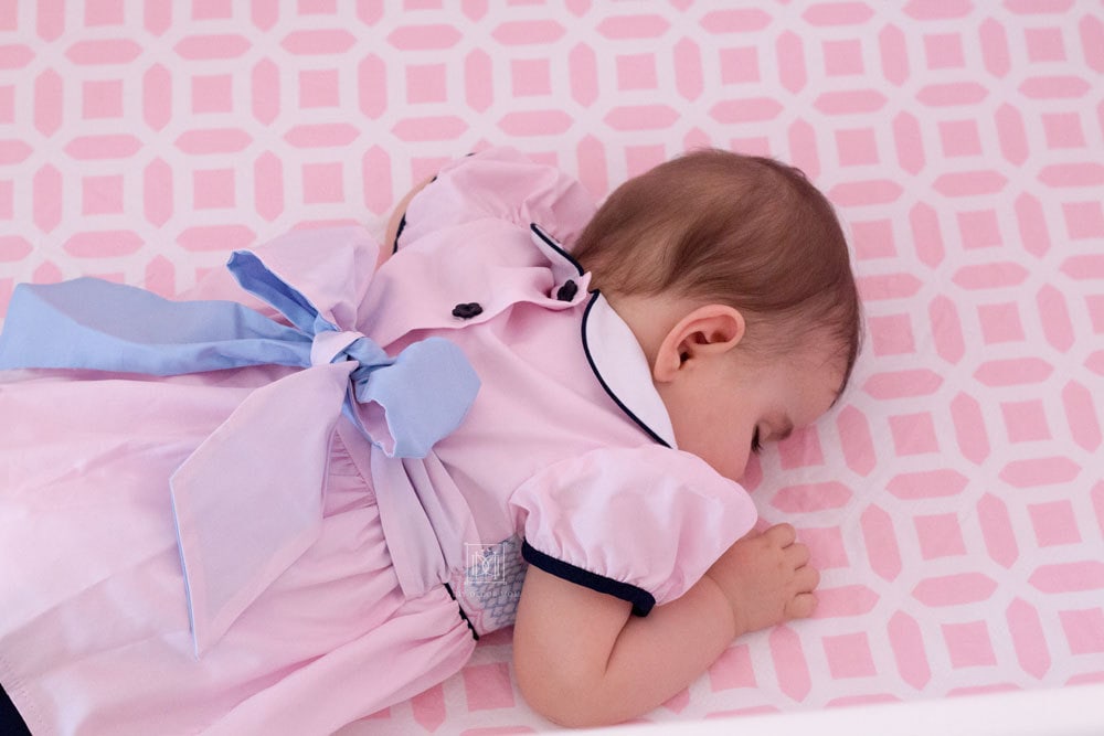 baby girl asleep in pink crib with smocked dress and blue bow