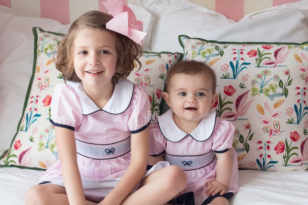 toddler and baby sister in pink smocked dresses on white bed in girls shared room