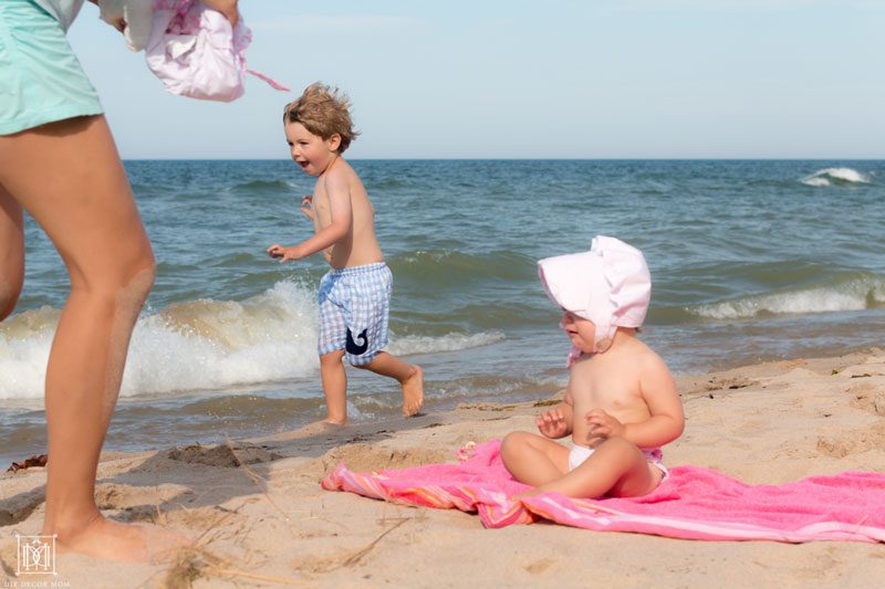 mom running to change baby's diaper and running toddler in the background at the beach