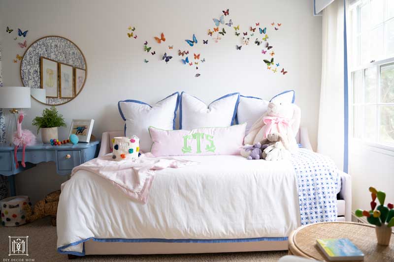 pink upholstered daybed in nursery with white bedding and monogrammed pillows- butterfly artwork wall decor