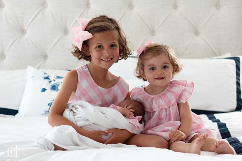 two girls in pink outfits sitting on white bed holding newborn