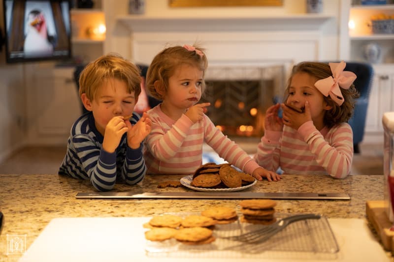 three siblings eating cookies at kitchen counter when one steals a cookie