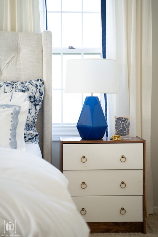 blue and white bedding in front of white curtains with modern bedside table and blue bedside table lamp