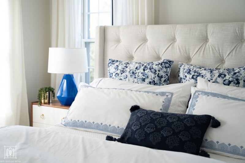 beautifully made blue and white bed with euro pillows on cream headboard, white curtains, and BM Classic Gray walls