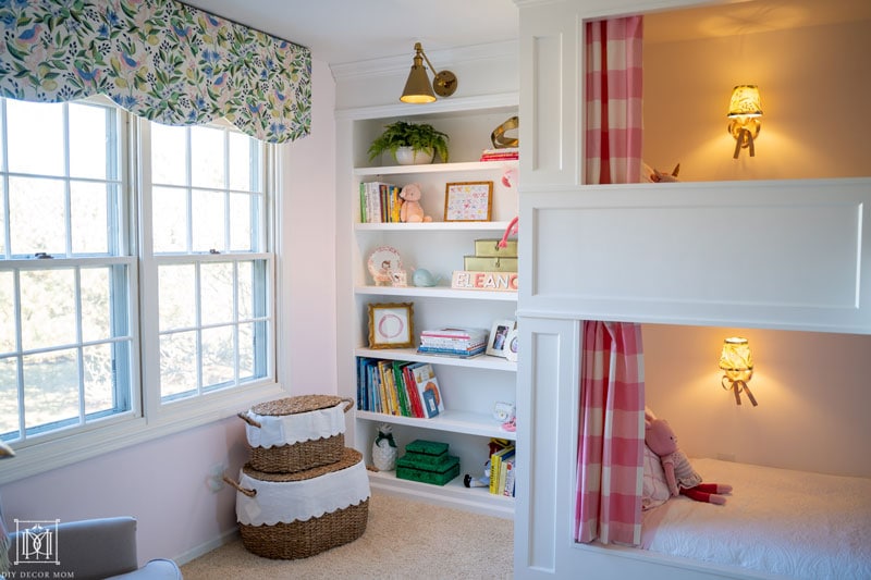 white bookcases with brass lighting in girls shared bunk room with toy storage in wicker baskets and scalloped pelmet box