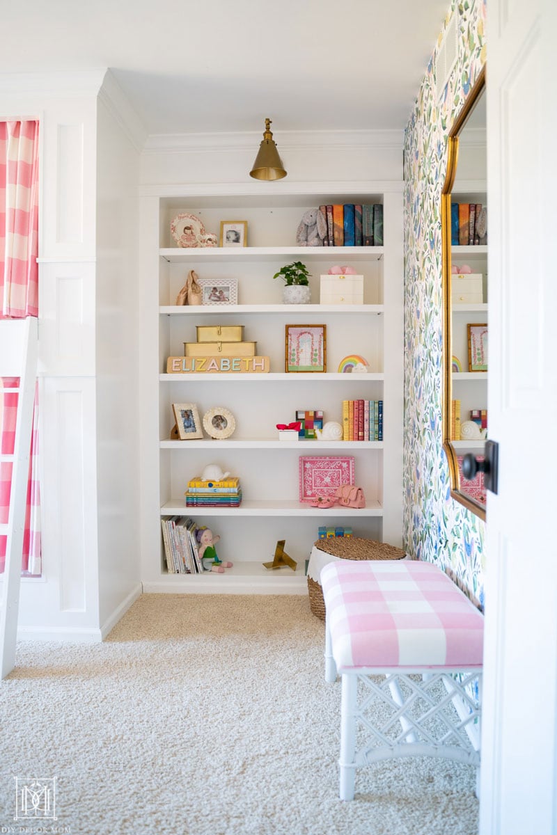 white built-in bookcases in girls bedroom with brass sconce and pink buffalo check design elements next to a green and white wallpapered wall